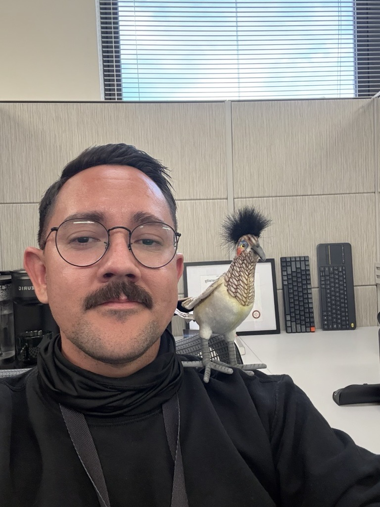 Luis Terrazas sitting at his cubicle with the roadrunner award on his shoulder.